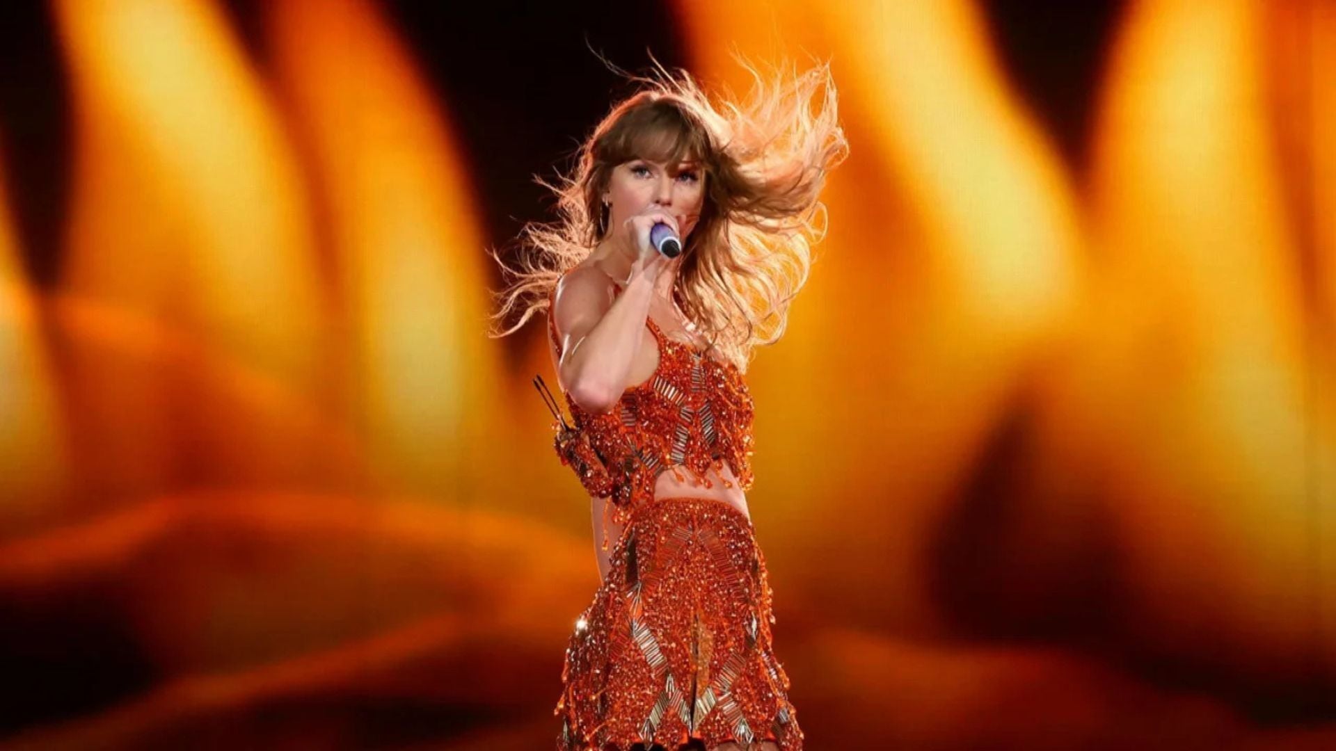 taylor swift vestida de naranja con un microfono con fondo naranja
